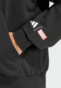 Schwarzer Sweatshirt mit gerippten Bündchen und Saum. Der linke Ärmel zeigt weiße Streifen und das rote "MARVEL"-Logo. Seitentaschen sind vorhanden.