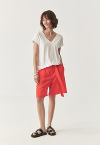 Witte V-hals t-shirt, lichte stof. Felrode linnen shorts met strik taille en zakken. Zwarte sandalen met hardware accenten.