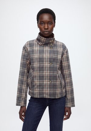 Femme portant une veste à carreaux grise et marron avec un col montant asymétrique boutonné et un jean bleu foncé, debout devant un fond uni.