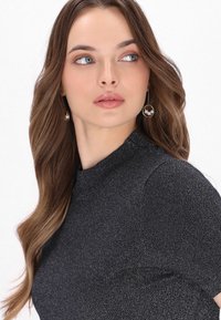 Top nero texturizzato con collo alto e maniche corte. Il modello indossa orecchini rotondi in oro con perle. Capelli lunghi e fluenti.