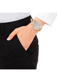 Goldfarbene Uhr mit einem runden, mit Strass besetzten Ziffernblatt, das funkelschöne Akzente zeigt, und einem verlinkten Armband, am Handgelenk getragen.