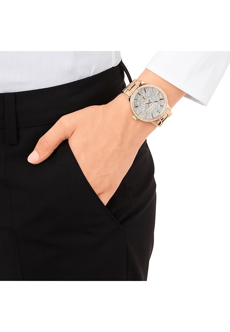 Goldfarbene Uhr mit einem runden, mit Strass besetzten Ziffernblatt, das funkelschöne Akzente zeigt, und einem verlinkten Armband, am Handgelenk getragen.