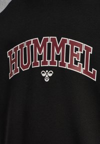 Schwarzer Pullover mit auffälligem dunkelrotem "HUMMEL"-Schriftzug, der über einem weißen Bienenlogo gewölbt ist. Weiches Material mit klassischem Rundhalsausschnitt.