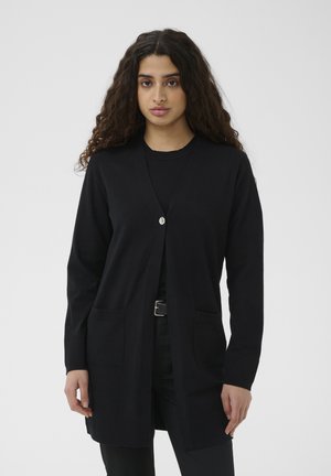 Jonge vrouw met lang krullend haar die een zwarte cardigan draagt met zakken aan de voorkant en één knoop, staand tegen een witte achtergrond.