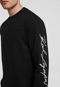 Hombre con sudadera negra de manga larga con el texto en cursiva blanca "Noble League" en la manga derecha y texto negro sutil en el pecho.
