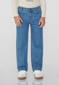 Světlé modré džíny s rovný střih, obsahující pět kapes, přední zapínání na knoflík a kontrastní šití. Vloženy s bílými teniskami.