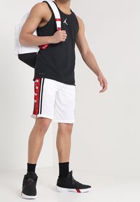 Débardeur noir, shorts blancs avec des bandes latérales rouges et baskets noires. Le sac à dos est blanc avec des accents noirs. Design athlétique simple.