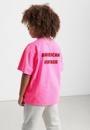 Enfant aux cheveux bouclés portant un t-shirt rose vif avec l'inscription "AMERICAN VINTAGE" dans le dos et un pantalon gris clair, debout à l'intérieur.