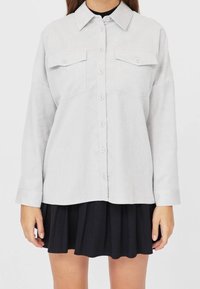 Lichtgrijs overhemd met kraag, twee borstzakken, lange mouwen en een ontspannen pasvorm, gecombineerd met een zwarte rok.