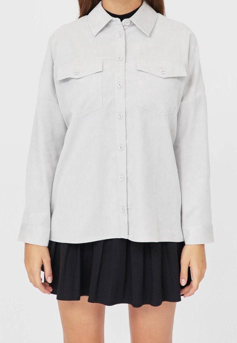 Lichtgrijs overhemd met kraag, twee borstzakken, lange mouwen en een ontspannen pasvorm, gecombineerd met een zwarte rok.