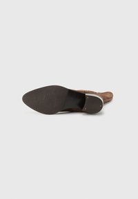 Chaussure à talons hauts en daim marron avec un bout pointu et une tige texturée. Semelle noire lisse avec une pointe arrondie et des accents matériels visibles.