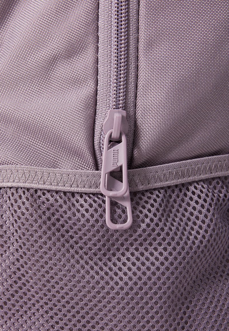 Bolsa de malla morada con una tela texturizada, que cuenta con un cierre de plástico y una lengüeta. El diseño incluye una sección inferior sólida en un color contrastante.