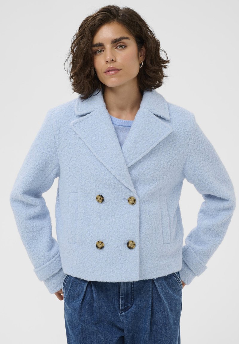 Veste bleu clair à double boutonnage en tissu texturé, présentant des boutons dorés, des revers crantés et des poches latérales.