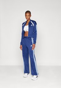 Adidas treningsdress i marineblå, med hvite tre striper på ermene og bena. Laget av glatt stoff, med en jakke med glidelås og bukser med elastisk liv.