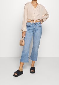 Henkilö, jolla on yllä vaaleanbeige lyhyt paita, joka on solmittu vyötäisiltä, siniset levenevät farkut ja mustat liukuvat kengät, pitää kädessään pientä käsilaukkua.