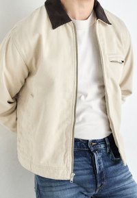 Hombre con chaqueta beige y cuello oscuro sobre camisa blanca y jeans azules, de pie con las manos detrás de la espalda.