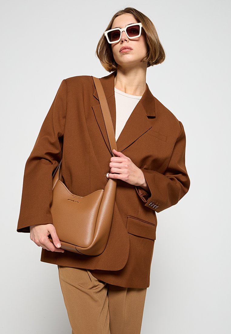 Bruine oversized blazer met een geknipt revers, gecombineerd met een beige top en tan kleurige broeken. Het model houdt een gestructureerde bruine leren handtas vast.