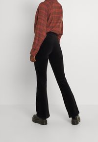 Svarta kordflorsbyxor med utställda ben och strukturerad textil, kombinerat med en röd och rutig långärmad skjorta. Utstyrseln framhäver en avslappnad passform.