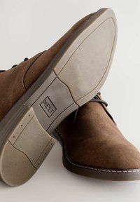 Brązowe zamszowe buty z czarnymi sznurówkami i gumową podeszwą. Podeszwa ma teksturowaną powierzchnię i widoczne logo marki na pięcie.
