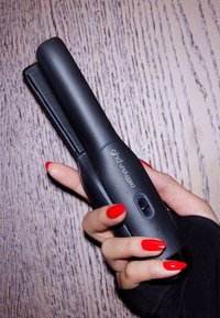 GHD noire tenue dans une main, avec une surface texturée, un bouton d'alimentation et un design élégant et minimaliste. Ongles rouges visibles.