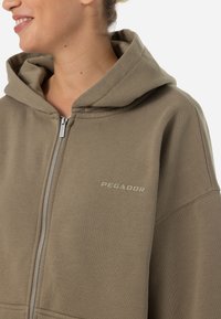 Sudadera con cremallera de color verde olivo, hecha de un material suave, que presenta una gran capucha, puños de canalé y un sutil logo "PEGADOR" en el pecho.