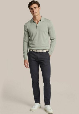 Man draagt een lichtgroen poloshirt met lange mouwen, zwarte broek, witte riem en witte sneakers, staand tegen een effen beige achtergrond.