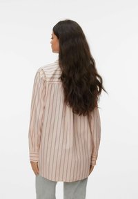 Femme aux cheveux longs et foncés portant une chemise ample rose pâle à rayures verticales et un pantalon gris clair, debout de dos sur un fond blanc.