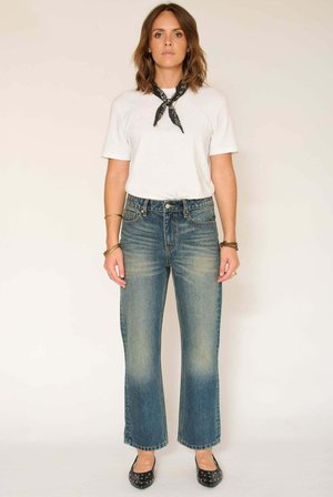 Jean en denim bleu taille haute avec jambes évasées, texture délavée, poches latérales et coutures contrastantes. Porté avec un t-shirt blanc et des ballerines noires.