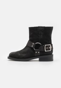 Botte de cheville en suède noir avec un talon plat, une large sangle avec une boucle en argent et un accent en anneau, et un bout arrondi. Ajustement réglable.
