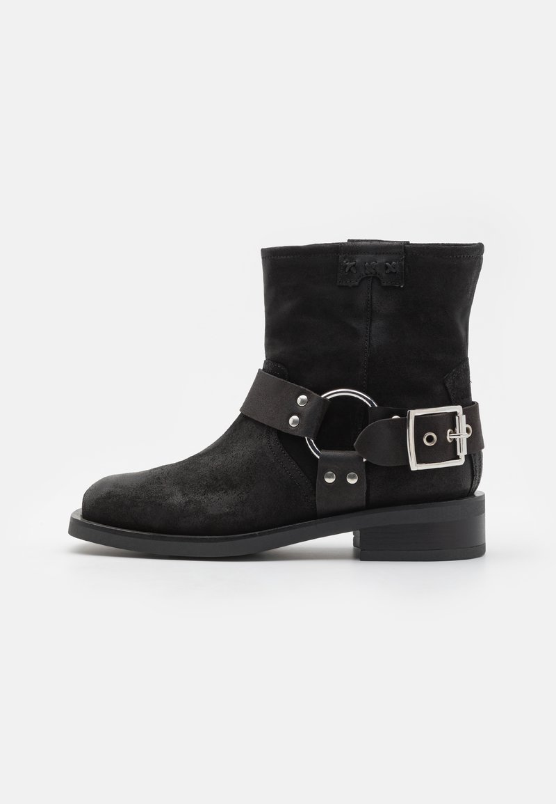 Botte de cheville en suède noir avec un talon plat, une large sangle avec une boucle en argent et un accent en anneau, et un bout arrondi. Ajustement réglable.