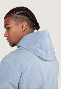 Helleblaue Kapuzenjacke mit einer knitterigen Textur, die ein schlichtes Design und eine verstellbare Kapuze aufweist. Der Stoff wirkt leicht und lässig.