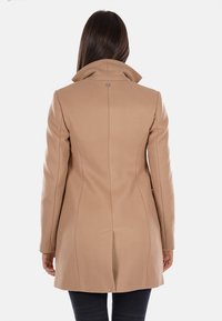 Cappotto beige su misura con colletto alto, vestibilità strutturata e dettagli di cucitura sul retro, realizzato in un tessuto liscio. Senza hardware visibile o tasche.