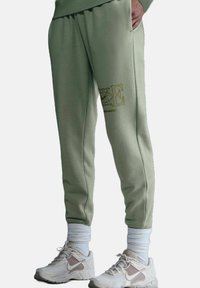 Pantaloni sportivi in cotone verde con cintura elastica, polsini a costine e dettaglio del logo Nike dorato sulla coscia sinistra. Abbinati a sneakers bianche.