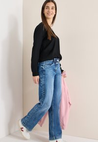 Femme portant un haut à manches longues noir, jean bleu, baskets blanches, marchant en tenant une veste rose devant un mur beige.