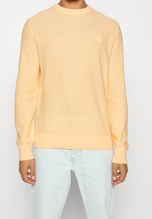 Mand iført en lys ferskenfarvet rundhalset sweater og lyseblå jeans, stående mod en ensfarvet baggrund med hænderne afslappet langs siderne.