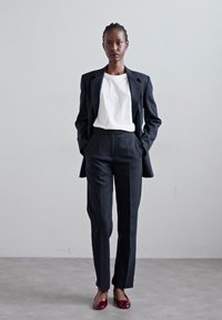 Costum negru, prelucrat, cu un sacou structurat și pantaloni drepți, asortat cu o cămașă albă și pantofi de culoare burgundă, pe un fundal simplu.