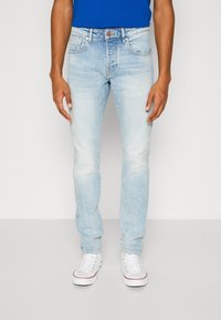 Jasnoniebieskie dżinsy o wąskim kroju, z efektami sprania, designem z pięcioma kieszeniami oraz brązowym zapięciem na guzik. Połączone z białymi trampkami.