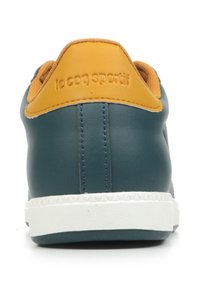 Teal leren sneaker met een gele kraag en een witte zool. Kenmerkt zich door subtiele stiksels en een geperst logo op de achterkant. Goede, gladde textuur.
