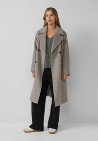 Vrouw in een lange beige dubbele-breasted jas, grijze V-hals trui, zwarte wijde broek en witte sneakers, staande tegen een effen achtergrond.