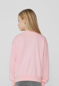 Růžová mikina vyrobená z měkké tkaniny, s volným střihem, dlouhými rukávy a žebrovanými manžetami a lemem. Nosená přes šedé džíny.