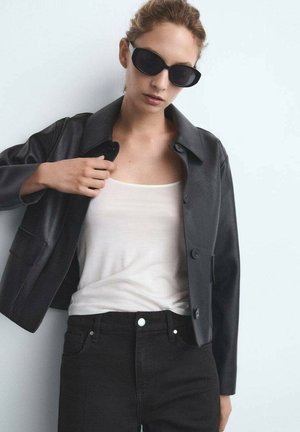 Chaqueta de cuero negra con cuello y cierre de botones, llevada sobre una camiseta sin mangas clara y jeans negros. El modelo lleva gafas de sol oscuras.