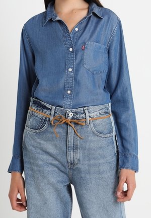 Kobieta ubrana w zapinaną na guziki niebieską koszulę dżinsową, włożoną do wysokich jasnoniebieskich dżinsów z cienkim brązowym paskiem zawiązanym na kokardę.