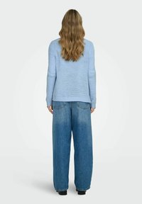 Woman with long wavy hair wearing a light blue knit sweater and wide-leg blue jeans, standing facing away against a plain background.