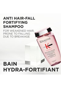 Champú Kérastase Genesis Bain Hydra-Fortifiant en una botella transparente rosa con tapa negra. Bloque geométrico transparente de fondo; la etiqueta incluye detalles del producto.