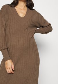 Donna che indossa un vestito a coste marrone con maniche lunghe e scollatura a V profonda, una mano sulla vita e l'altra rilassata lungo il fianco.