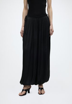 Žena oblečená v dlhej čiernej plisovanej sukni so šnúrkou v páse a čiernych sandáloch na opätku stojaca pred jednofarebným svetlým pozadím.