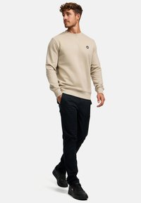 Beiger Hoodie mit gerippten Bündchen und Rundhalsausschnitt, ausgestattet mit einem kleinen schwarzen Logo, kombiniert mit schwarzen Hosen und schwarzen Sneakers.