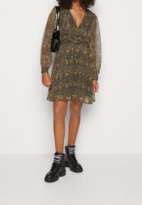 Robe à manches longues avec un décolleté en V profond, ornée d'un motif abstrait multicolore dans des nuances de marron, de bleu sarcelle et d'orange ; associée à des bottes noires à lacets.