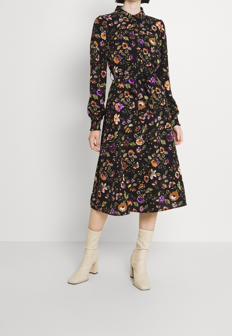Femme portant une robe midi noire à fleurs à manches longues avec une ceinture à la taille et des bottines beiges à talons, debout sur un fond blanc.