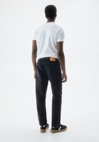 Jeans de mezclilla negra con corte recto, con un parche de cuero marrón en la parte trasera, combinados con una camiseta blanca y zapatillas negras.